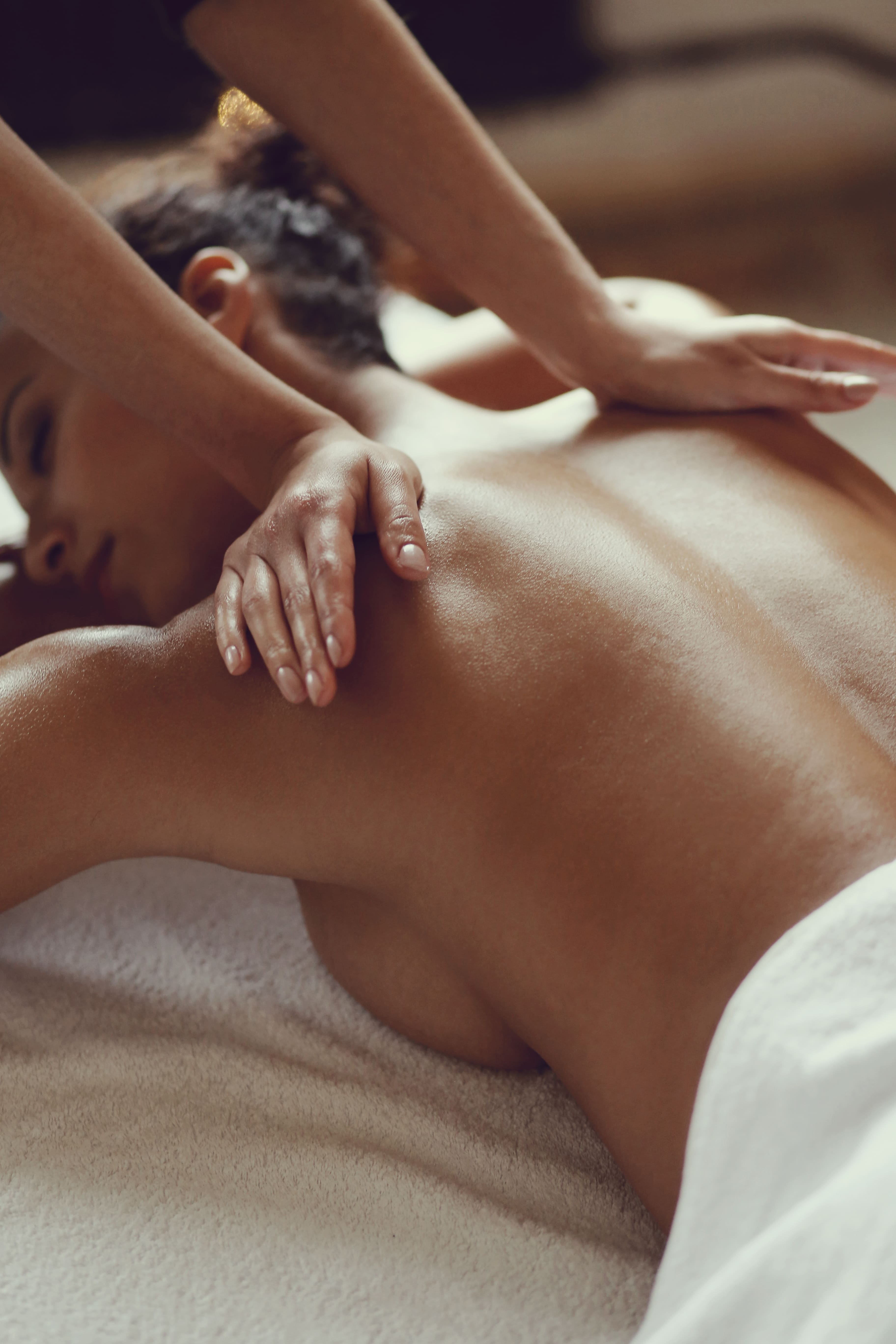 A person is receiving a relaxing back massage on a white towel, with dim lighting creating a calm, soothing atmosphere. Skin glistens under soft light.